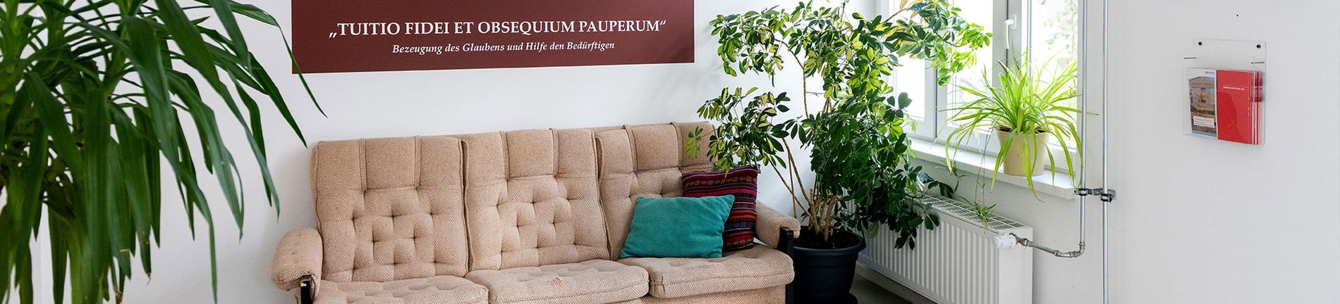 Zu sehen ist ein beiges Sofa an einer Wand, an der ein großes Bild hängt mit dem Malteserkreuz und dem Leitsatz: Bezeugung des Glaubens und Hilfe den Bedürftigen