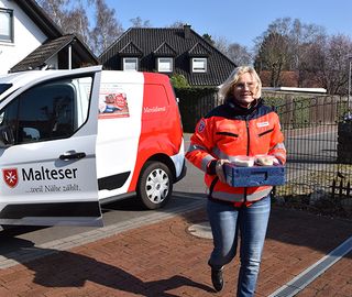 Menüservice: Eine Frau in Malteser Dienstbekleidung mit einer Wärmebox vor einem Malteser Fahrzeug
