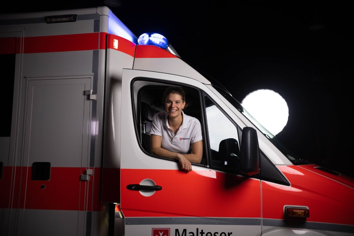 Eine junge Notfallsanitäterin schaut lächelnd aus dem Beifahrerfenster eines Rettungswagens.