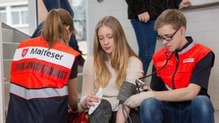 Ein Schüler und eine Schülerin in Malteser-Einsatzbekleidung messen den Blutdruck einer Schülerin in einem Schultrakt.