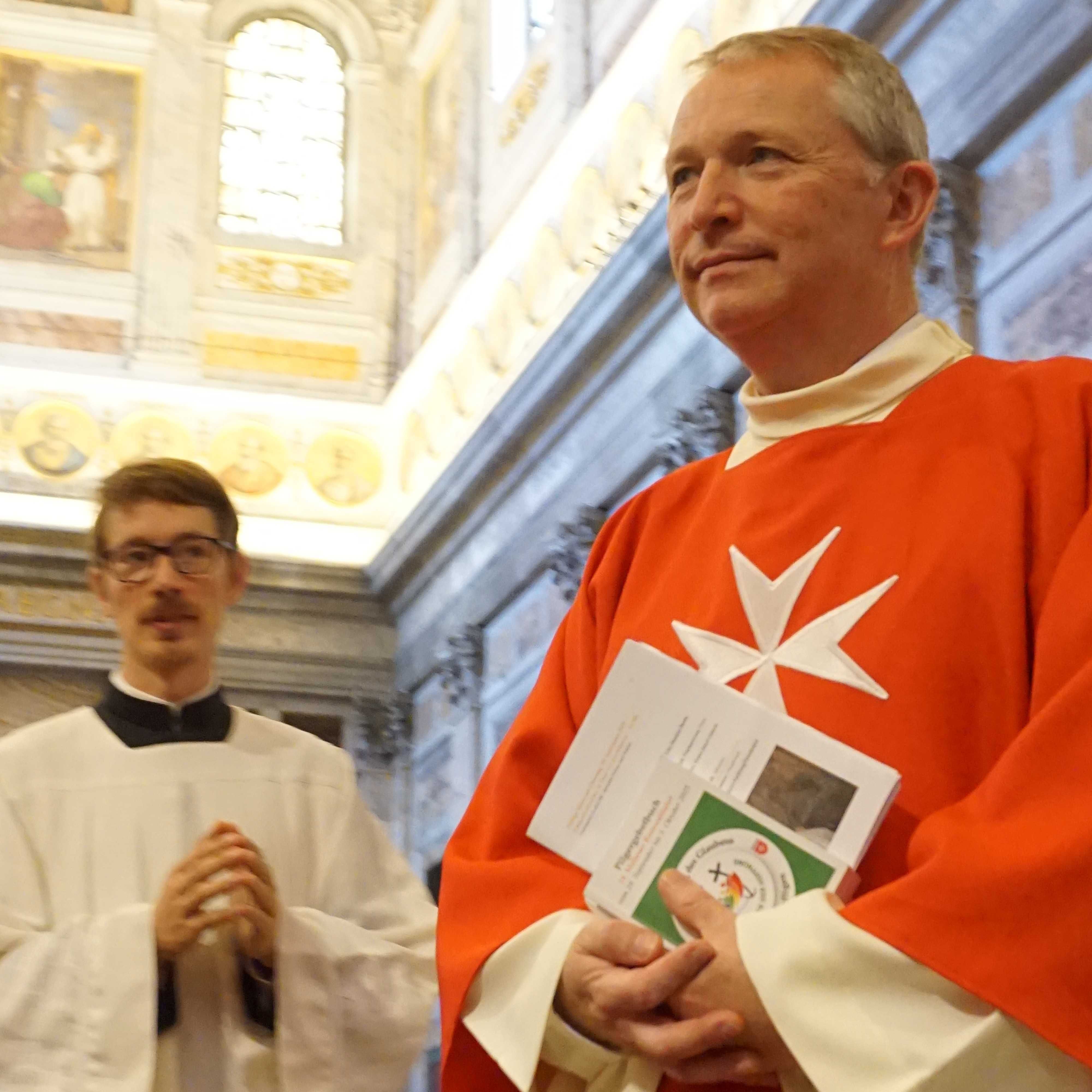 Zwei Geistliche in Kirchengewändern, einer in rotem Gewand mit weißem Malteserkreuz, der andere in weißem Gewand, stehen in einer Kirche mit bemalten Wänden und hohen Fenstern.
