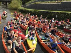 14 Kanus auf dem Wasser, in denen jeweils mehrere Personen sitzen und freudig in die Kamera lächeln.