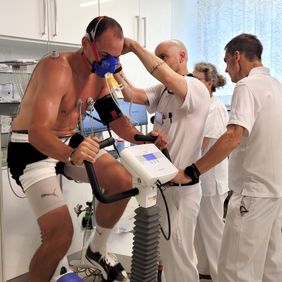 Leistungstest auf dem Ergometer mit Atemmaske, betreut von drei medizinischen Fachkräften in weißer Kleidung.