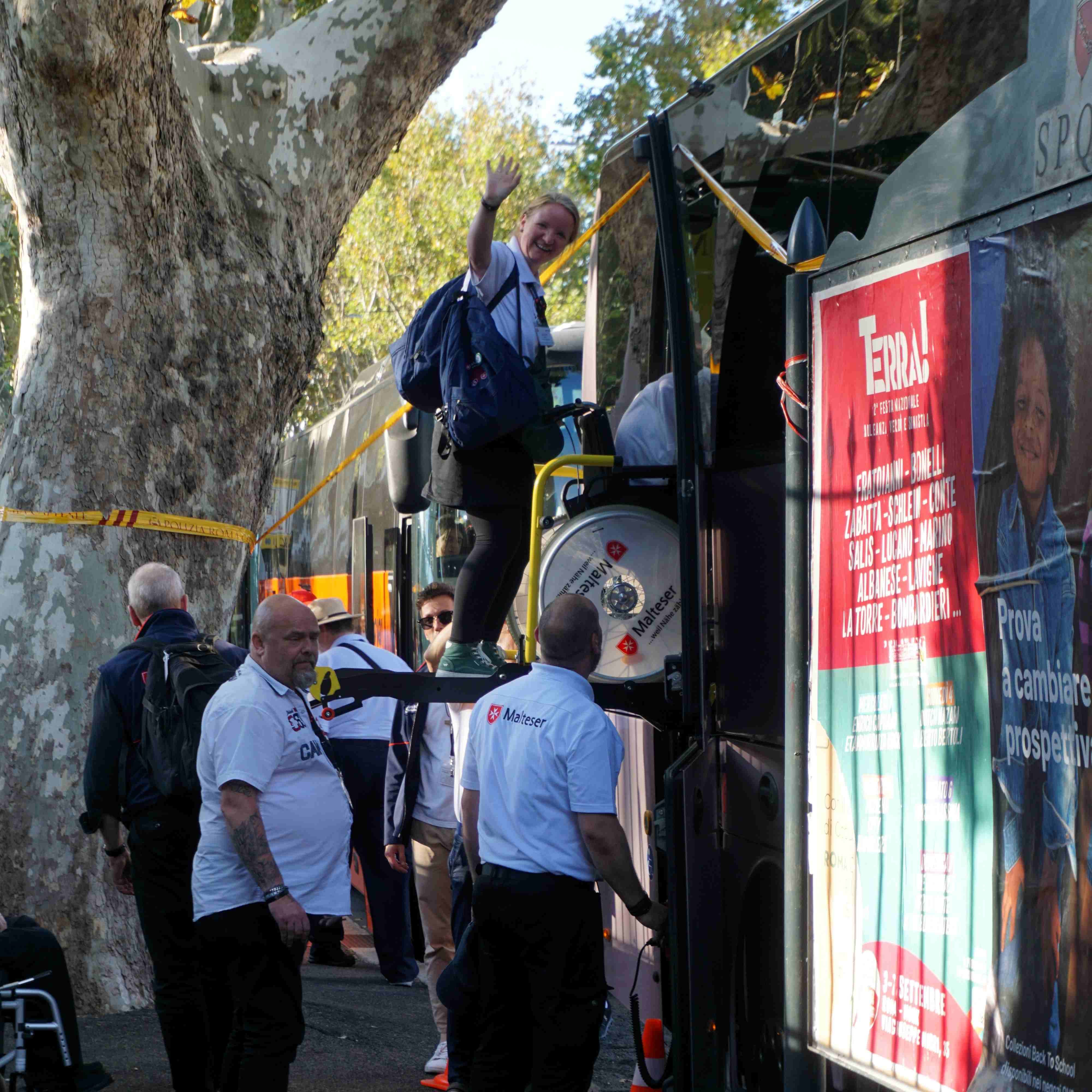 Person mit Rucksack steigt in Bus ein, weitere Personen warten davor, Baum und Bus mit rotem Werbeplakat sichtbar.