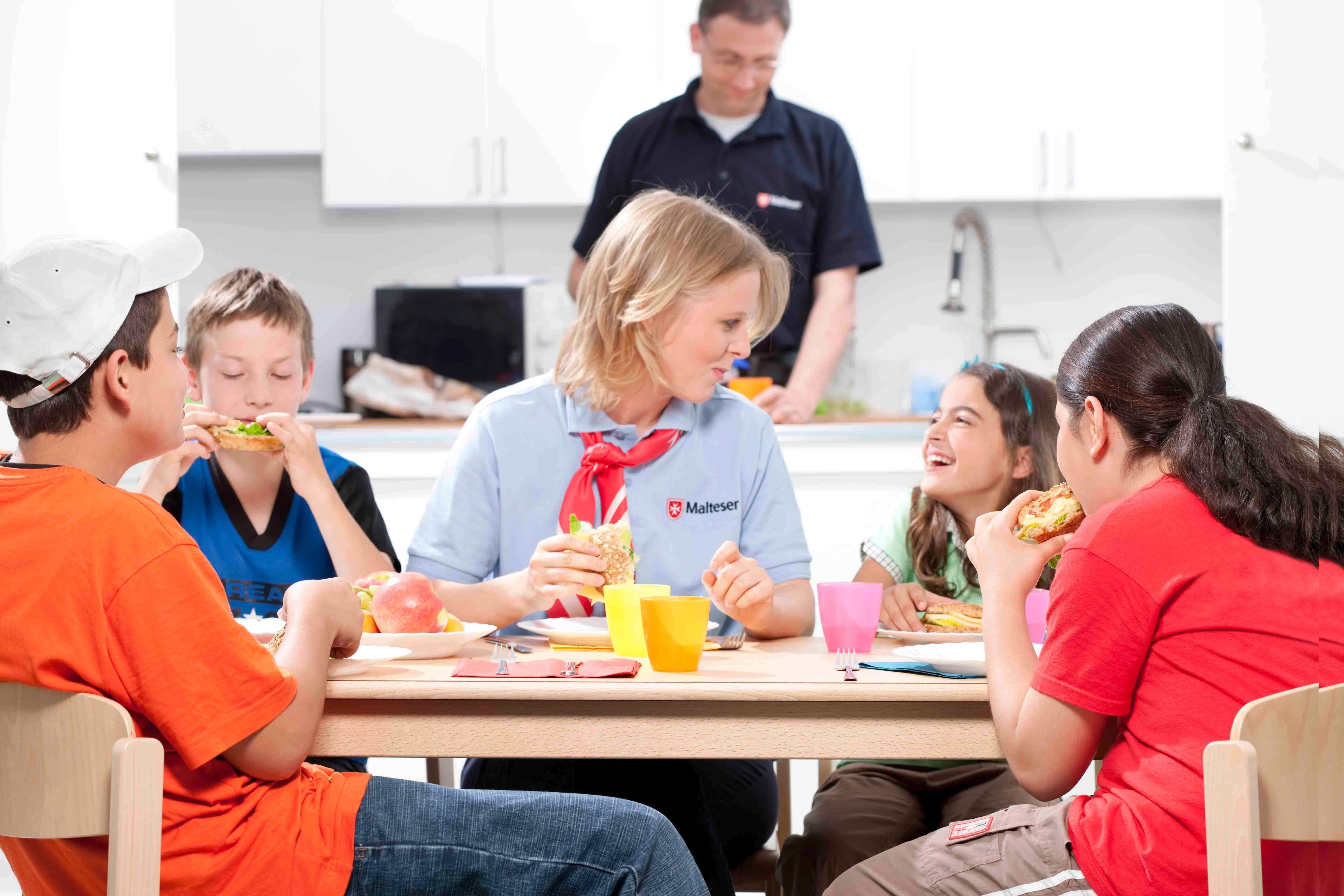 Ehrenamtliche Helferin isst mit Kindern am Tisch.