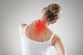 Frau im weißen Top fasst sich mit einer Hand an den Nacken und mit der anderen an den unteren Rücken.