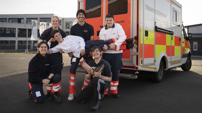 Sechs Jugendliche posieren in Malteser Kleidung und mit einem "abgerissenen Fuß" vor einem Rettungswagenu