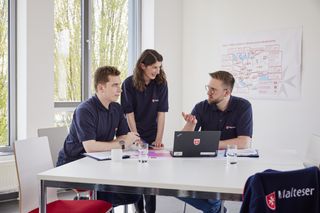 2 Malteser sitzen an einem Tisch. Eine Malteserin steht zwischen den beiden anderen. Sie beraten sich.