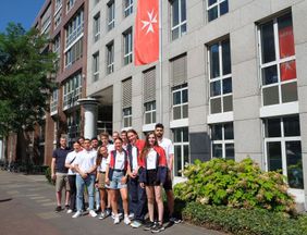 Gruppenfoto mit dreizehn jungen Menschen in Malteser Bekleidung vor der Malteser Fahne im Kaltenbornweg 3