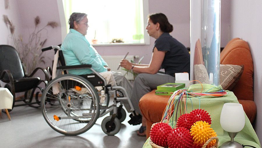 Eine Frau sitzt auf dem Sofa einer Frau im Rollstuhl gegenüber und zeigt ihr etwas auf dem Tablet