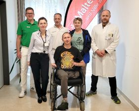 Patient im Rollstuhl mit sechs medizinischen Fachkräften in Klinikflur.