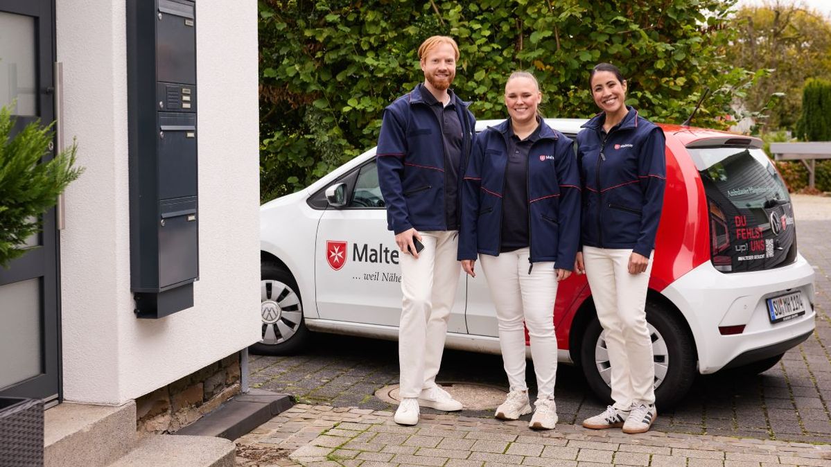 Ein Mitarbeiter und zwei Mitarbeiterinnen der ambulanten Pflege der Malteser stehen vor einem Auto.