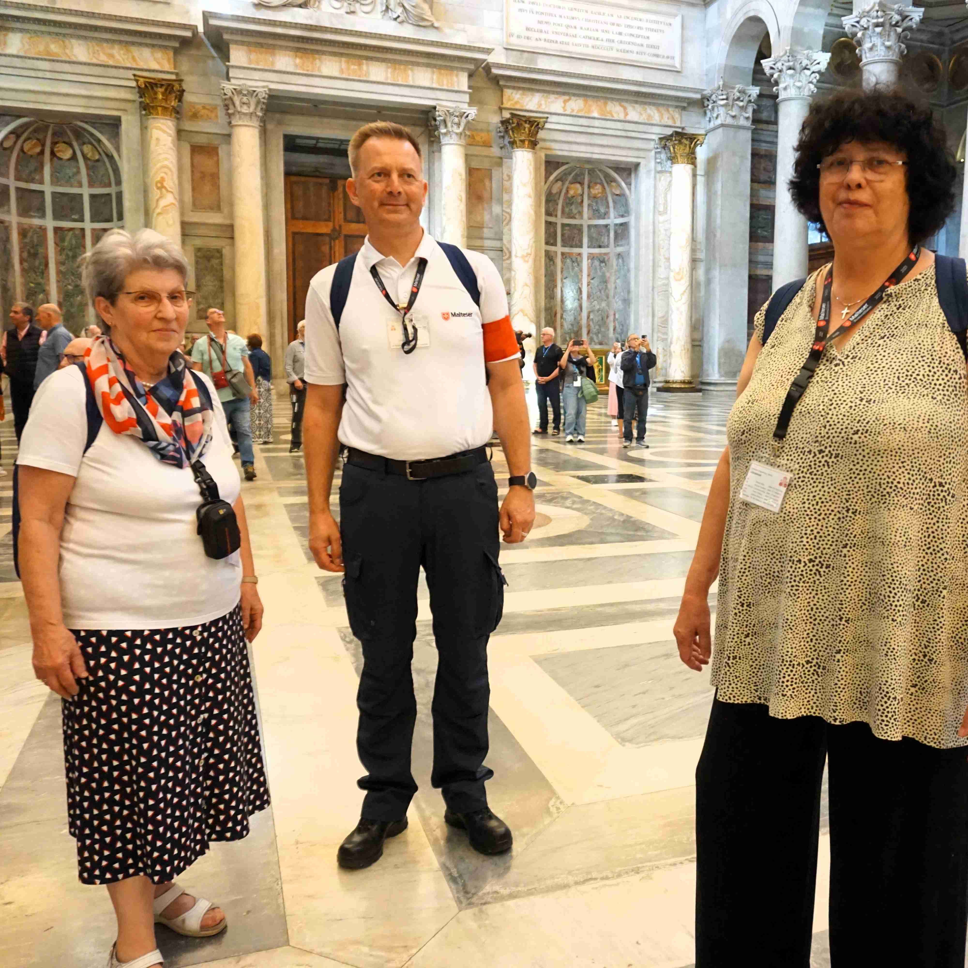 Drei Personen in einem großen, hellen Raum mit Marmorboden und hohen Fenstern, zwei Frauen und ein Mann, alle mit Ausweisen um den Hals.