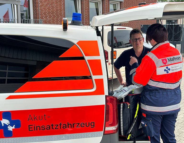 Zwei Personen in Malteser Einsatzkleidung stehen am geöffneten Heck eines Malteser Ensatzfahzeugs.