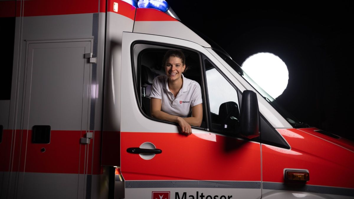 Eine junge Notfallsanitäterin schaut lächelnd aus dem Beifahrerfenster eines Rettungswagens.