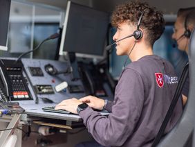 Ein junger Mann in einem Pullover, auf dem das Wort "Malteser" steht, sitzt in einem Fahrzeug vor einer Tastatur und einem Monitoren.