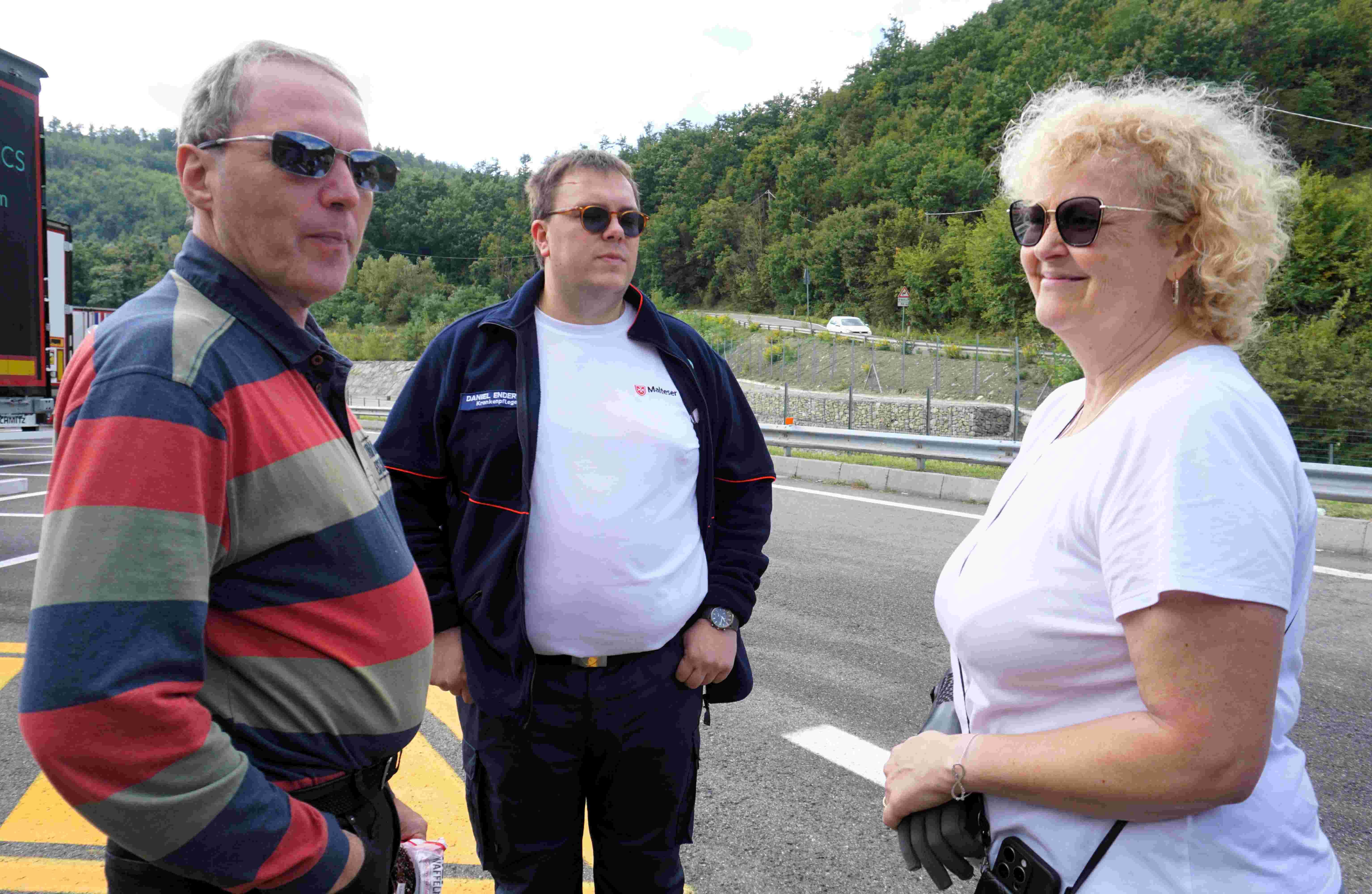 Drei Personen stehen auf einer Straße, umgeben von bewaldeten Hügeln, eine trägt ein rot-grau gestreiftes Hemd, eine ein weißes T-Shirt, eine eine dunkle Jacke.