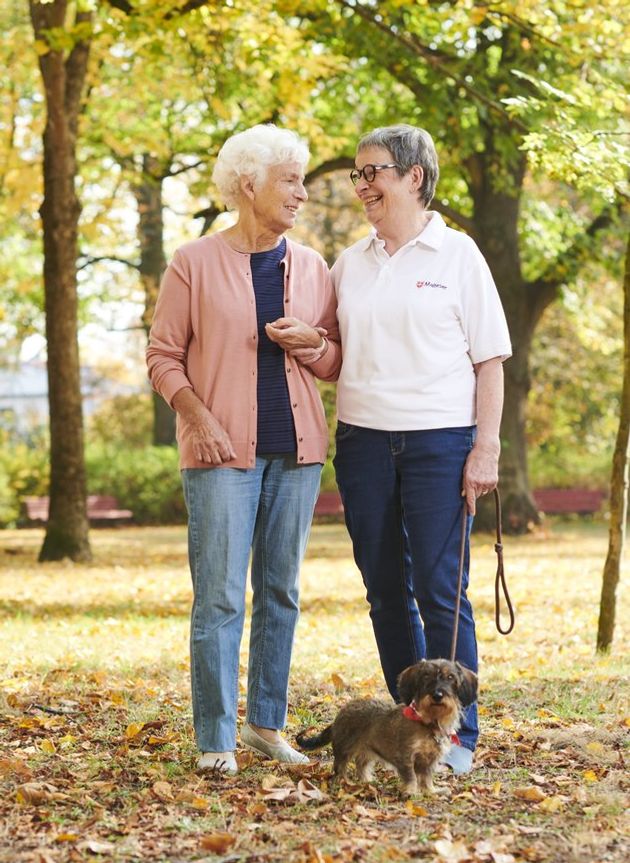 Eine ältere Dame wird von einer Ehrenamtlichen des Malteser Hilfsdienstes bei einem Spaziergang mit einem Dackel im Wald begleitet. Beide Frauen unterhalten sich und genießen die gemeinsame Zeit.