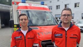 Zwei Männer in Rettungsdienstbekleidung stehen vor einem Rettungswagen.