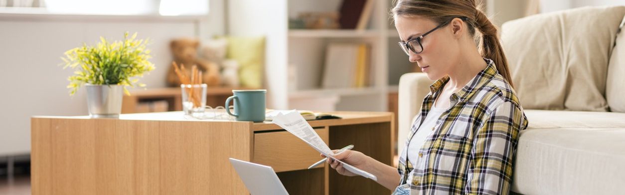 Junge Frau mit Brille sitzt auf dem Boden mit Laptop, Stift und einem Zettel.