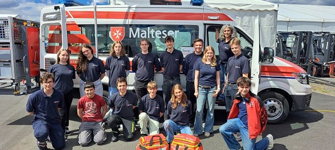 14 Schulsanis stehen oder knien gemeinsam vor einem Malteser Rettungswagen und schauen lächelnd in die Kamera. 