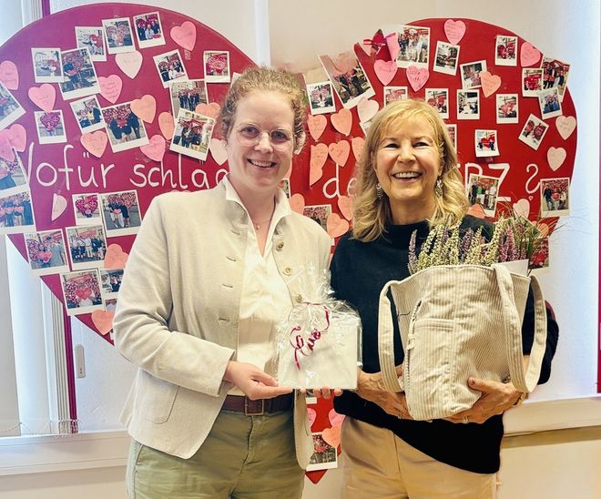 Zwei Frauen stehen vor einem roten Herz, in den Händen halten sie Geschenke