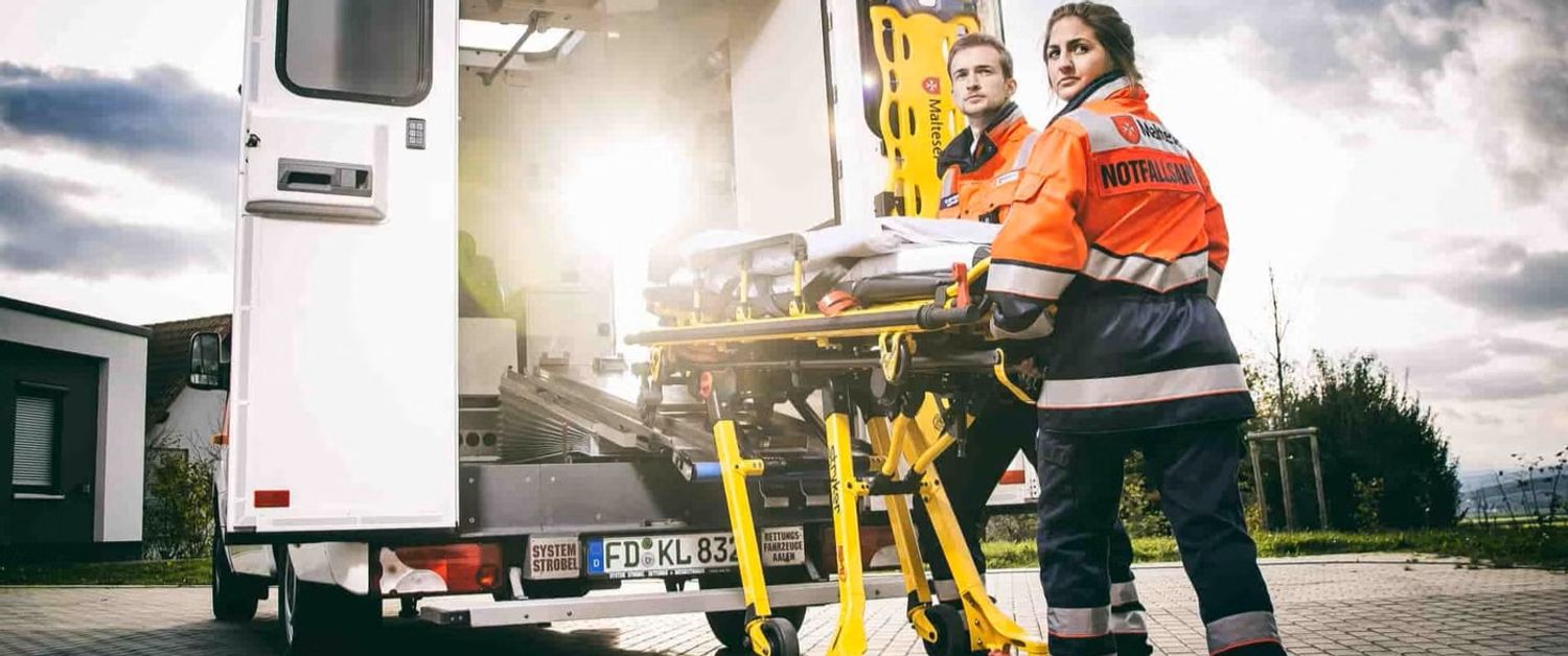 Malteser Rettungsdienstmitarbeitende verladen Krankenliege in Rettungswagen.