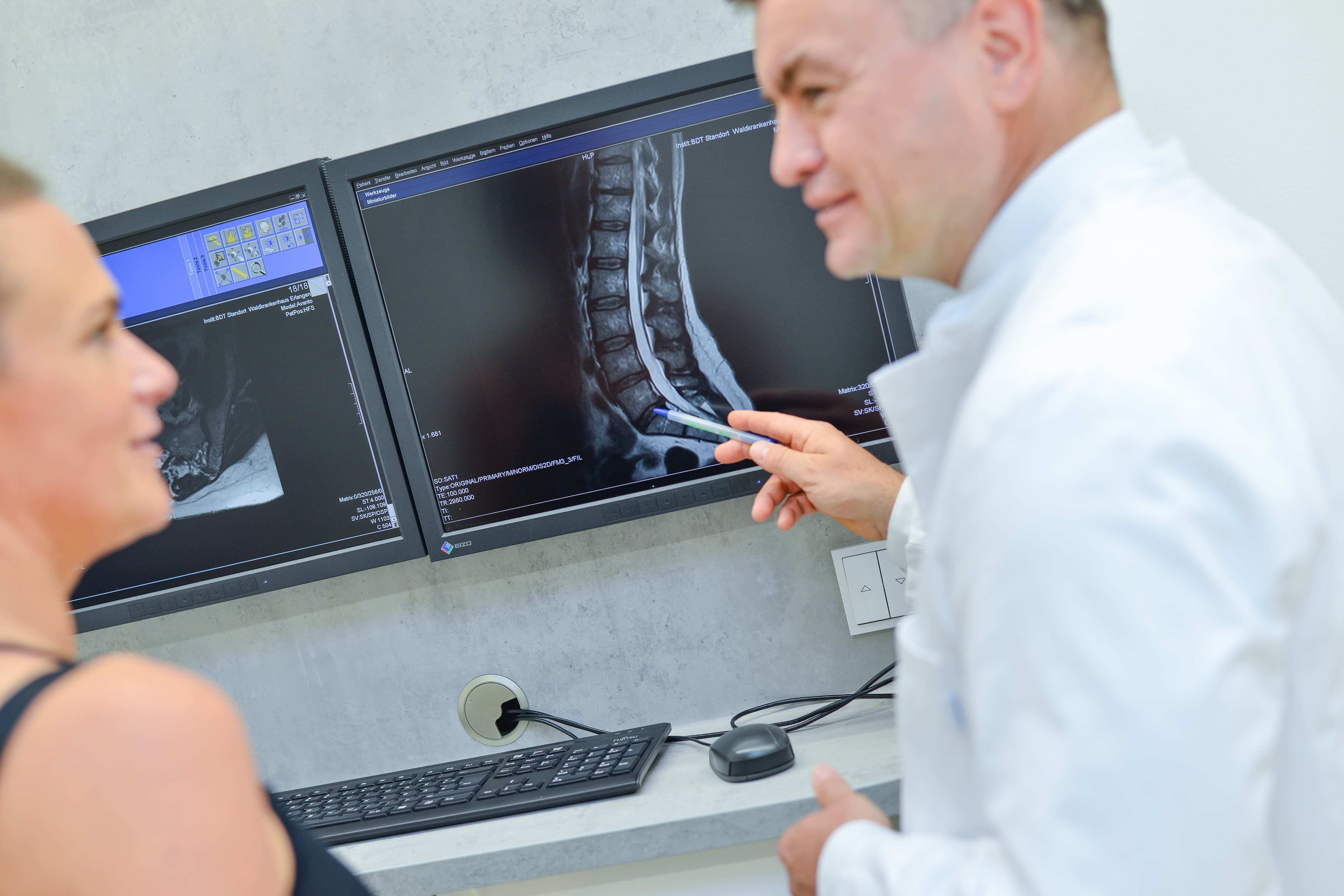 Ärztliche Fachkraft bespricht mit einer Patientin die MRT-Aufnahme der Wirbelsäule an zwei Monitoren im Untersuchungsraum.