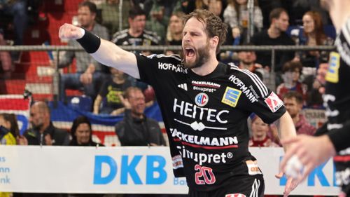 Handballspieler jubelt mit erhobener Faust auf dem Spielfeld, weitere Personen im Hinter-grund.