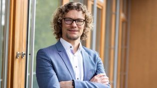 Businessporträt einer Person mit lockigem Haar und Brille im Anzug vor großen Fenstern.
