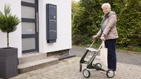 Eine ältere Frau mit Rollator und Mobilnotrufuhr am Handgelenk steht vor einem Hauseingang.