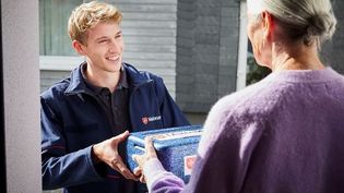 Ein junger Mann vom Malteser Menüservice übergibt einer älteren Dame ihr Essen in der Warmehaltebox an der Tür. 