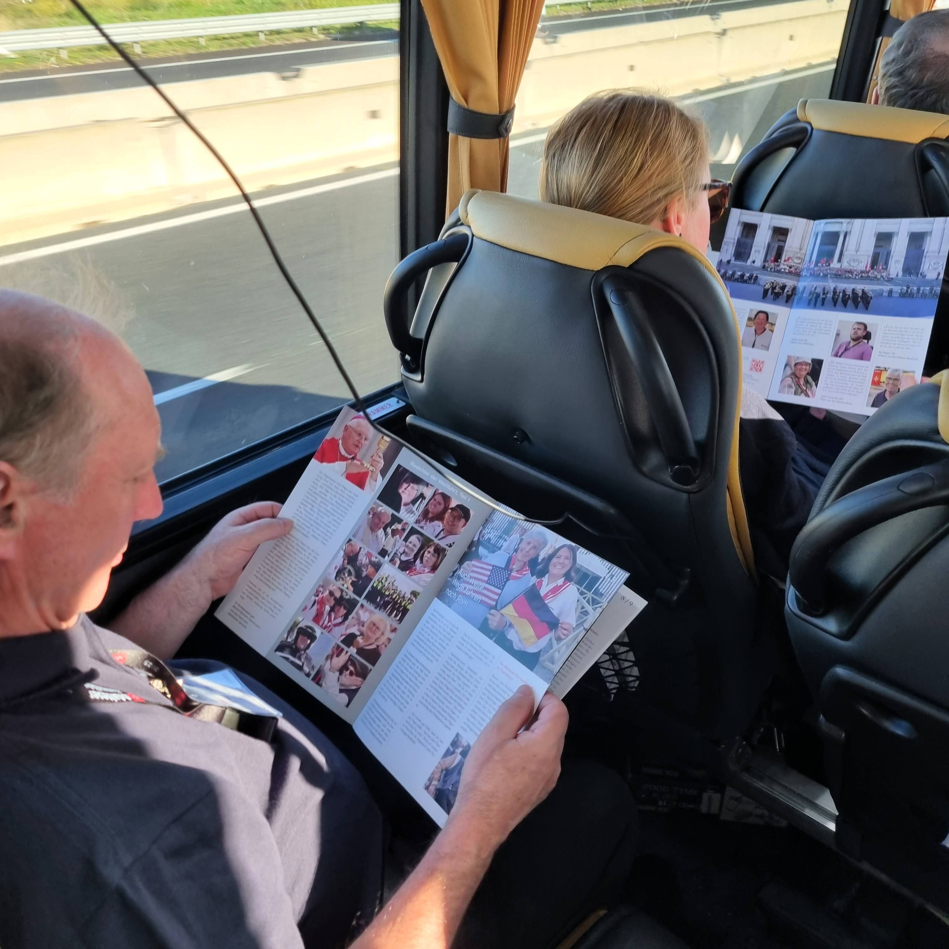 Person sitzt in einem Bus und liest eine Zeitschrift mit Bildern und Texten, weitere Person vor ihr ebenfalls lesend.