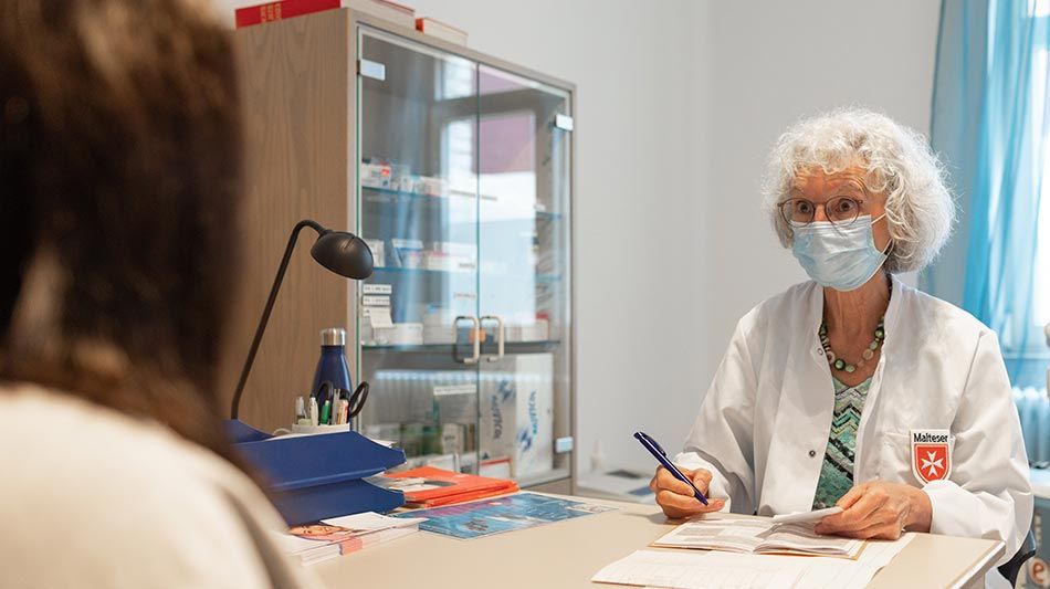 Eine Ärztin mit Mundschutz sitzt an einem Schreibtisch und spricht mit einer Frau