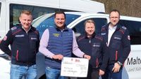 Gruppenfoto der an der Übergabe des symbolischen Spendenschecks beteiligten Personen vor einem Fahrzeug der Malteser