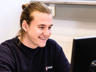 Junger Mann mit Malteser Pullover sitzt vor einem Laptop