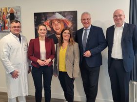 Fünf Personen posieren vor Wand mit Porträtfoto, darunter ein Arzt und mehrere Personen in Businesskleidung.