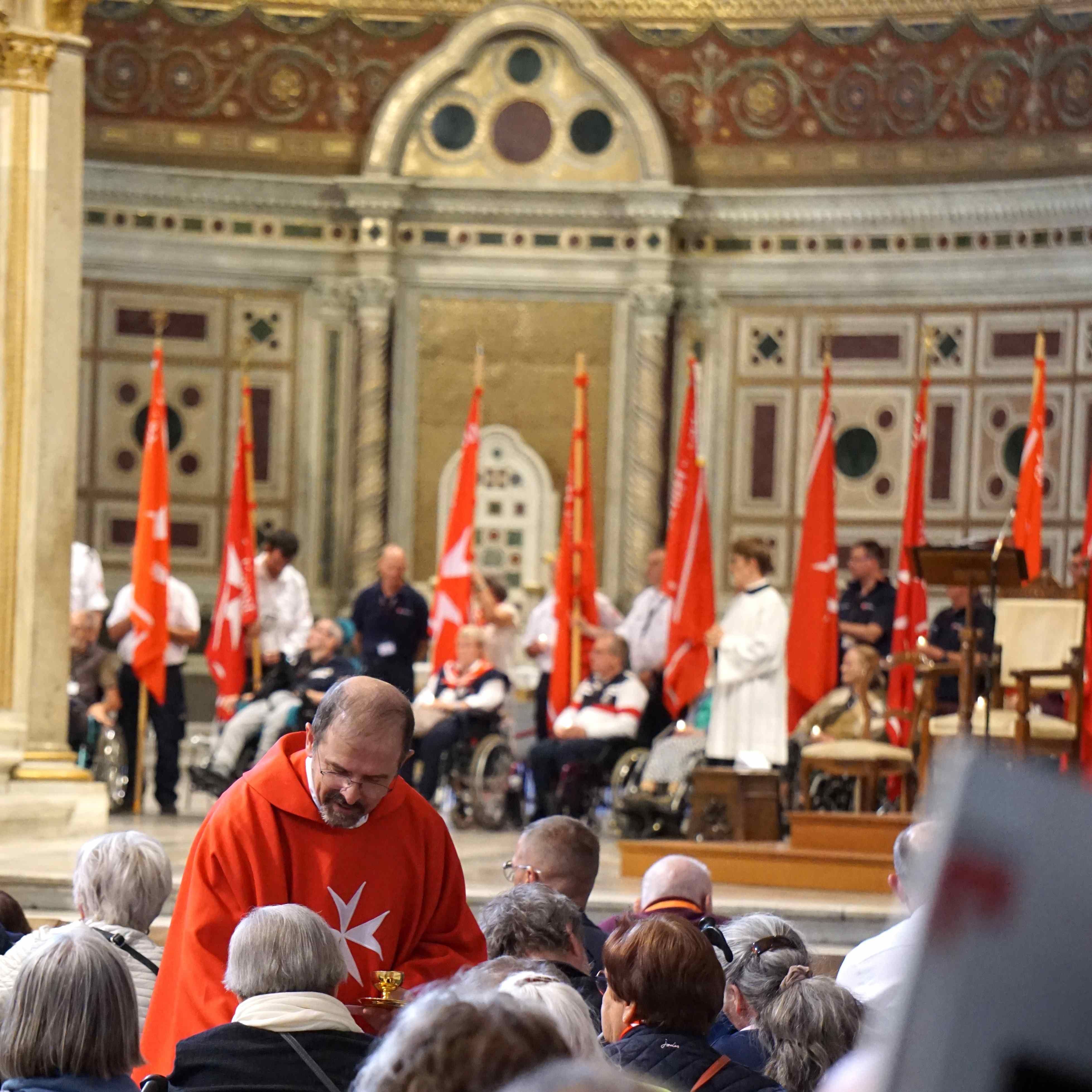 Pfarrer steht in einer Kirche im roten Gewand und beugt sich vor einen Menschen. Im Hintergrund sind weitere Personen und rote Fahnen zu sehen.