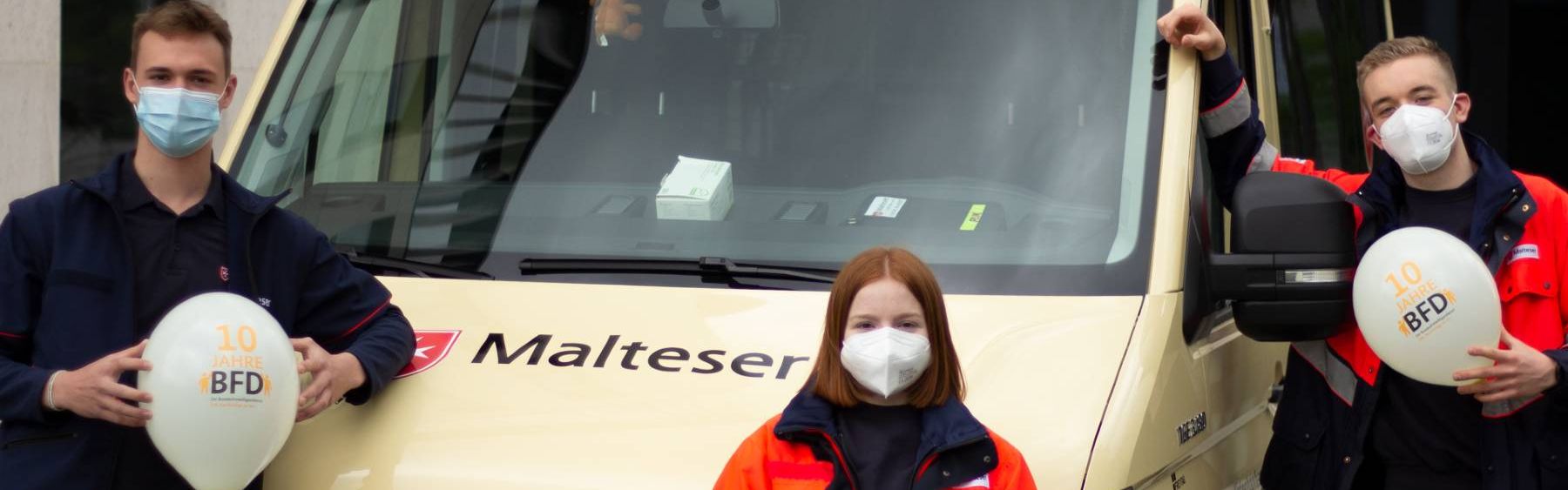 Eine junge Frau und zwei junge Männer halten Luftballons vor einem Kleinbus in die Höhe