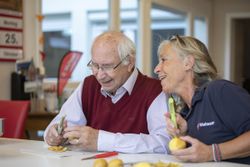 Ein älterer Mann und eine etwas jüngere Frau sitzen zusammen am Tisch und schälen Kartoffeln. Die Frau trägt ein blaues Malteser Shirt.