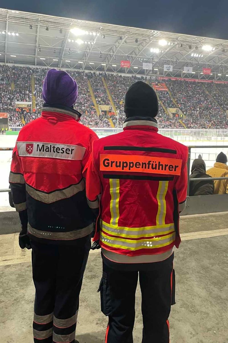 Rückenansicht zweier Malteser Einsatzkräfte in einem Eishockeystadion. 