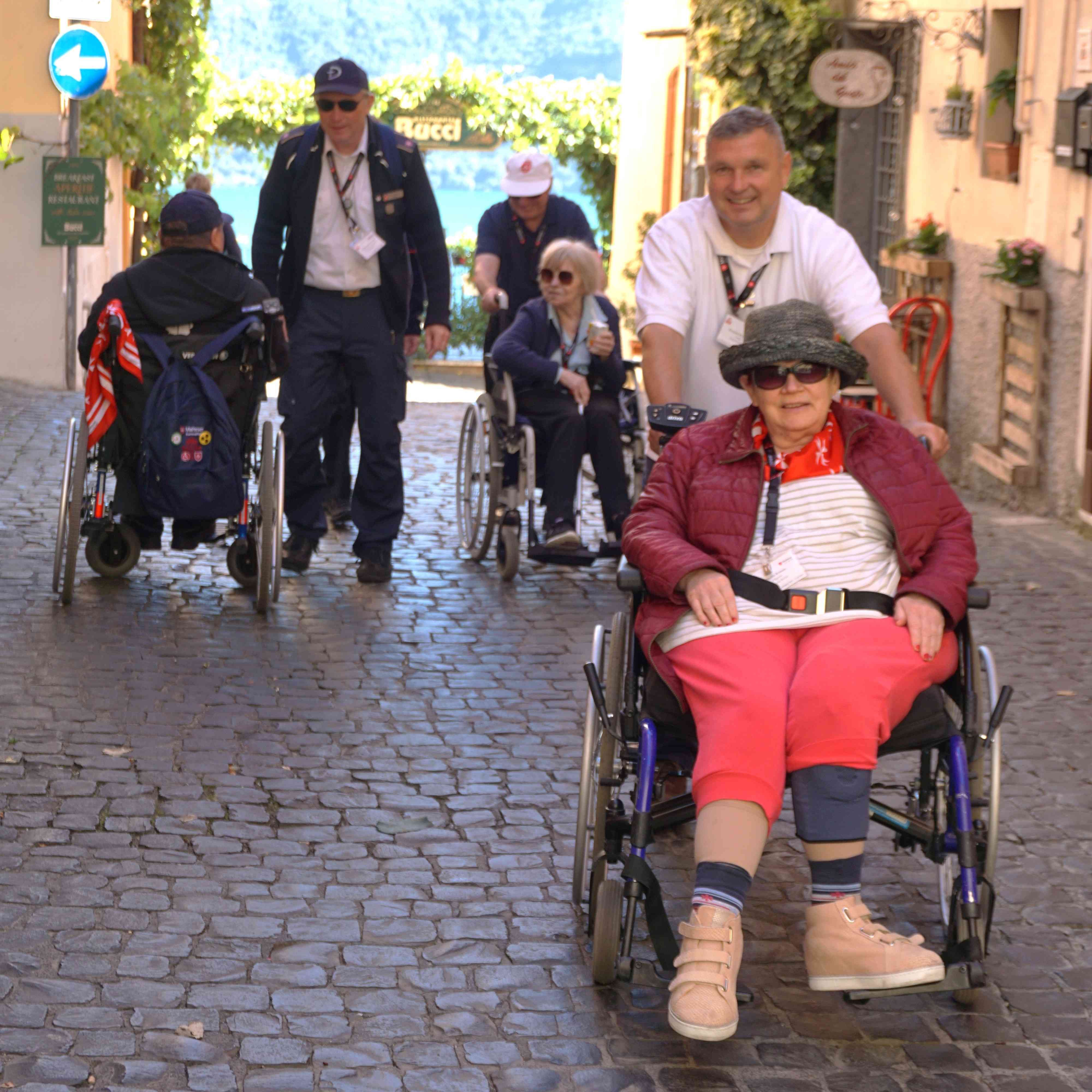 Menschen in Rollstühlen und Begleitpersonen gehen auf gepflastertem Weg in Castel Gandolfo, umgeben von Gebäuden und Pflanzen.