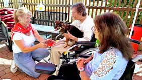 Eine Frau besucht mit einem Hund ältere Menschen. Die älteren Menschen freuen sich über den Besuch und streicheln den Hund.