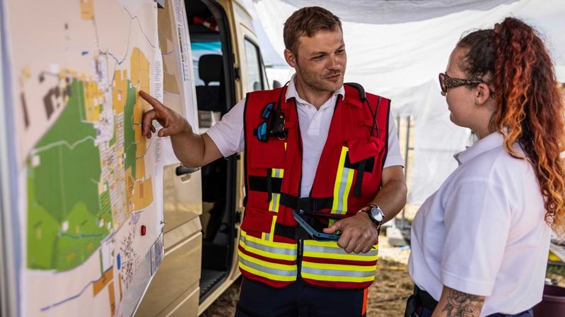 Ein Mann in einer roten Weste mit Reflexionsstreifen deutet auf eine Geländekarte und redet mit einer jungen Frau in einem weißen Hemd. 