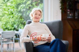 Eine Seniorin genießt ihren Kaffee zuhause und trägt ein Notrufarmband.