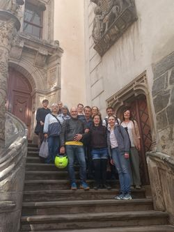 Eine Gruppe von Teilnehmenden steht auf den Stufen eines reich verzierten historischen Gebäudes und posiert für ein gemeinsames Erinnerungsfoto. Die kunstvoll gestaltete Fassade und die Treppenanlage verleihen der Szene einen besonderen Rahmen.