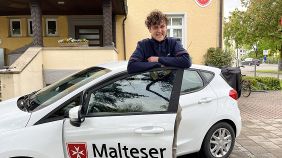Ein junger Mann in Malteser-Kleidung steht in der Tür eines Malteser-Autos.