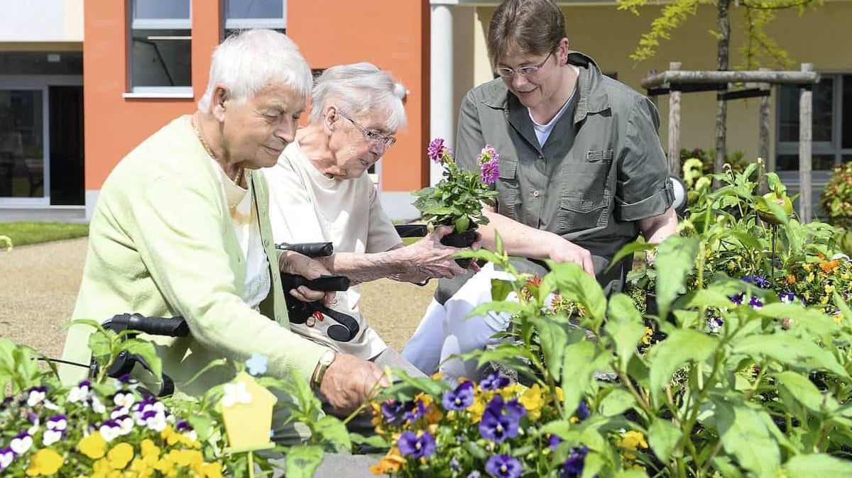 Zwei ältere Frauen und eine jüngere Frau bepflanzen ein Blumenbeet vor einem Gebäude.