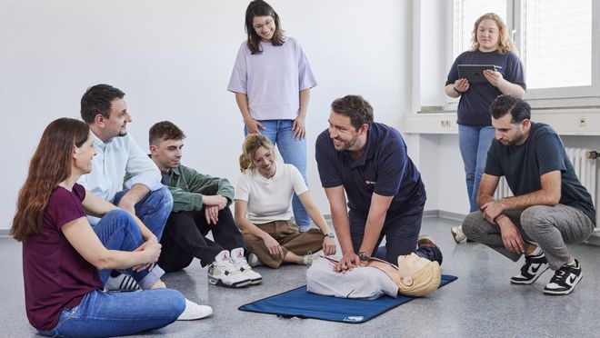 Ein Malteser Ausbilder demonstriert an einer auf dem Boden liegenden Übungspuppe die Herzdruckmassage, um ihn herum sitzen und stehen Kursteilnehmende