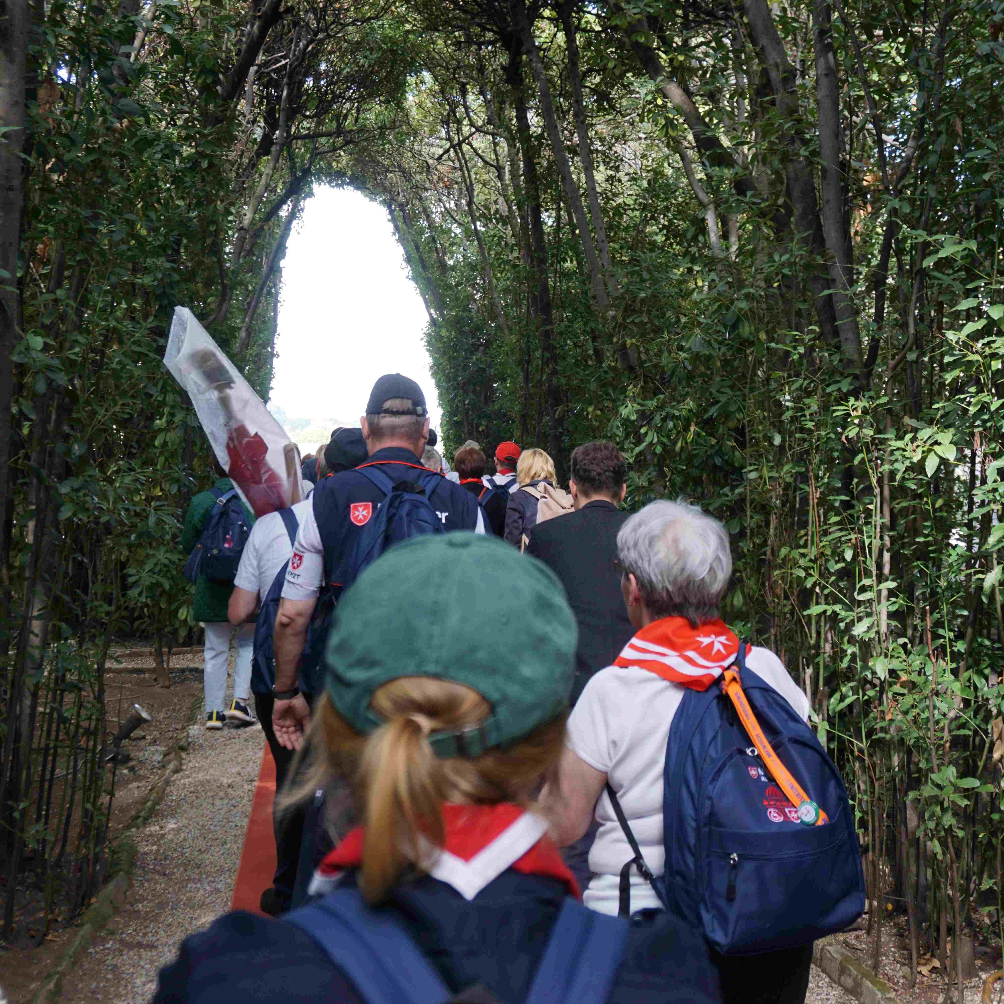 Menschen mit Rucksäcken und Halstüchern gehen auf einem Weg, umgeben von Bäumen.